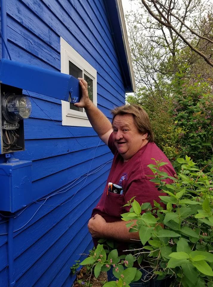 Tim Williams smiling beside a blue-painted house, his hand on the conduit above an electric meter