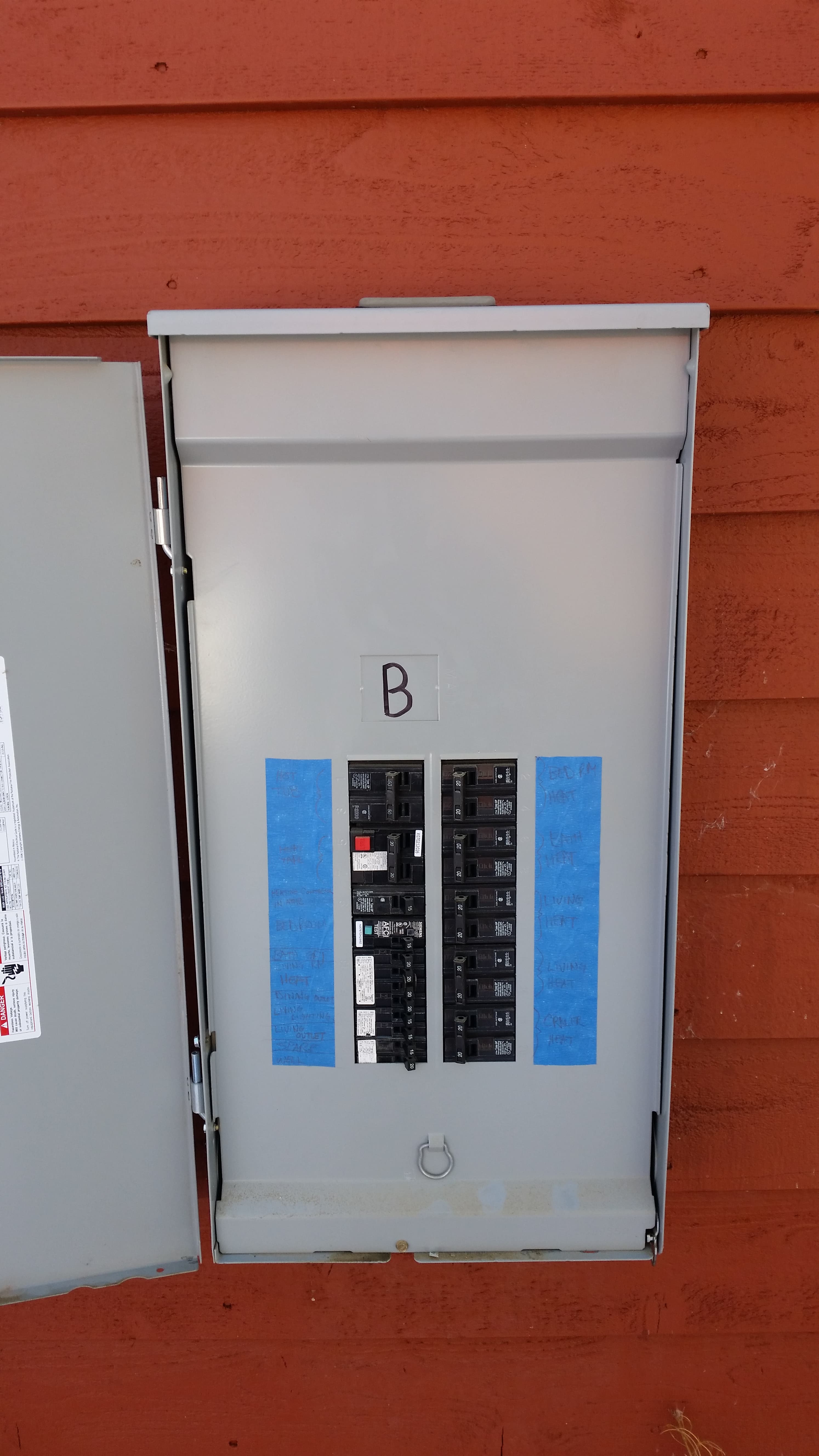 Outdoor breaker panel marked B on red siding, its circuits labeled on blue tape